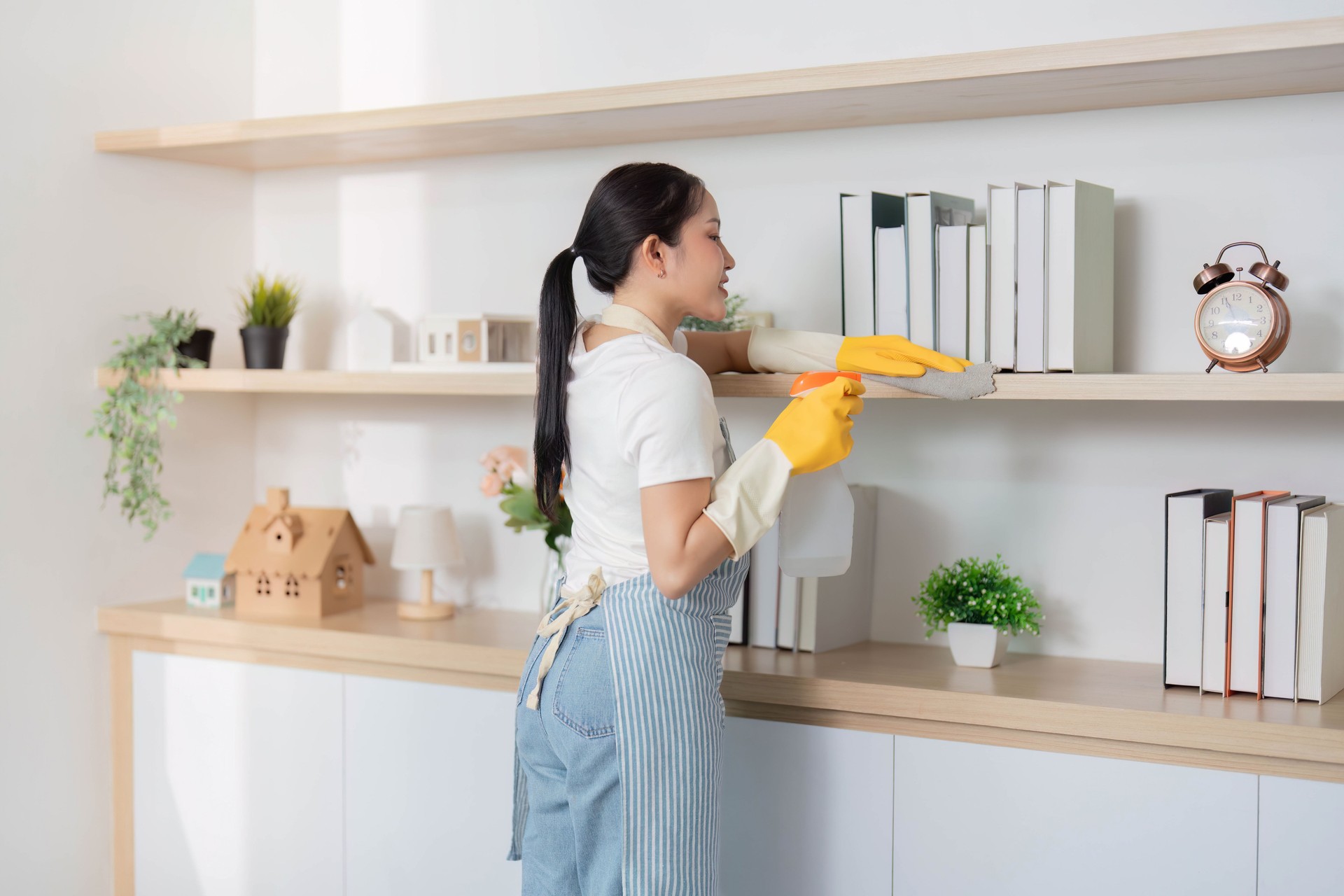 Home Cleaning. Young woman dusting shelves in a modern living room, promoting cleanliness and organization.